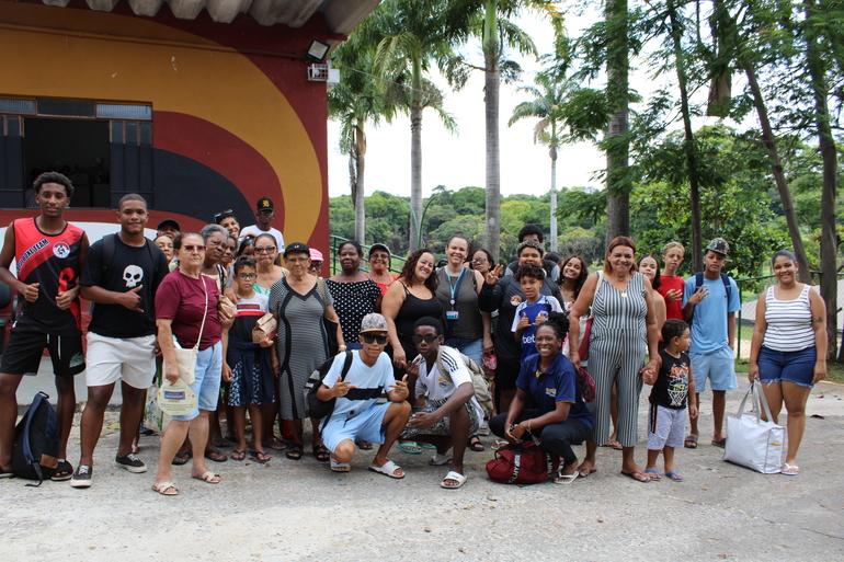 Em uma área externa arborizada, cerca de 30 pessoas de diferentes idades posam para uma foto em grupo. Elas estão reunidas em frente a um edifício com fachada em tons de vermelho, laranja e preto, enquanto, ao fundo, há diversas palmeiras e árvores de grande porte, compondo um ambiente verde e aberto. A maioria sorri para a câmera; alguns adolescentes fazem gestos com as mãos e duas pessoas estão agachadas na parte da frente do grupo.