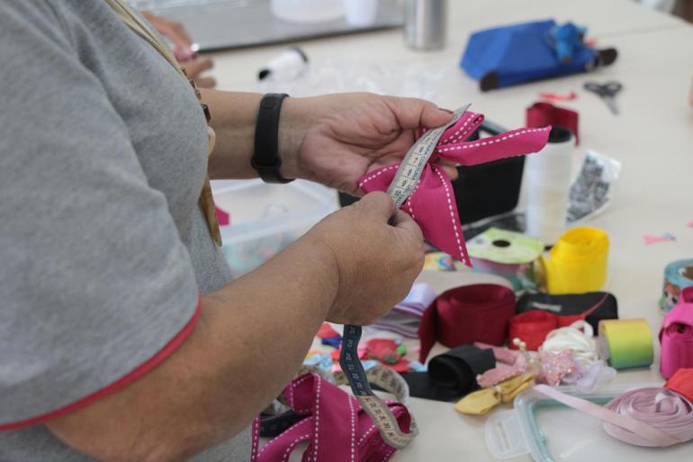 Pessoa confeccionando um laço de fita, utilizando materiais de artesanato em uma mesa de trabalho.