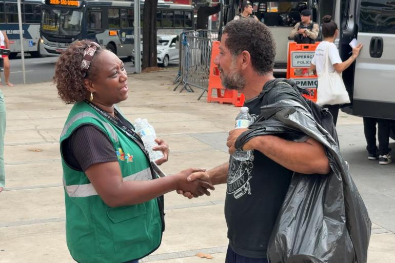 A foto mostra uma cena ao ar livre, em uma calçada movimentada de uma área urbana. No primeiro plano, duas pessoas conversam e apertam as mãos. À esquerda, uma mulher usa colete verde com faixas refletivas e segura uma garrafa de água. À direita, um homem veste camiseta preta e parece estar usando uma capa ou plástico escuro sobre os ombros; ele também segura uma garrafa de água. Ao fundo, há ônibus, veículos, agentes uniformizados, grades de isolamento e equipamentos de sinalização.