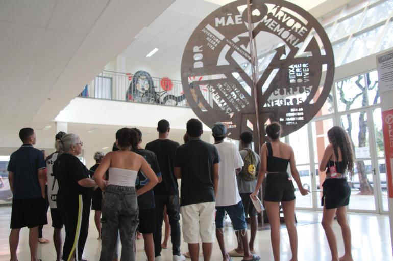 Programação Kilombo Aldeia reúne dança, capoeira e cultura afro no Centro de Referência das Juventudes Em ambiente interno amplo e iluminado, um grupo de jovens e adultos, vistos de costas, observa uma grande escultura metálica em formato de árvore. A obra ocupa o centro da imagem e traz palavras e expressões recortadas em sua estrutura, entre elas “mãe”, “memória” e “juventude negra viva”. Ao fundo, há mezanino, paredes claras e grandes janelas de vidro, que permitem a entrada de luz natural.
