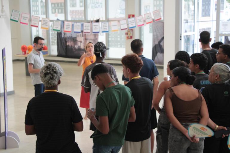 Em um espaço amplo e iluminado, um grupo de jovens está reunido em pé, de costas ou de lado para a câmera, acompanhando a fala de duas pessoas à frente. Ao fundo, há painéis expositivos, cartazes coloridos pendurados em um varal e grandes janelas de vidro. Algumas pessoas seguram materiais impressos nas mãos.