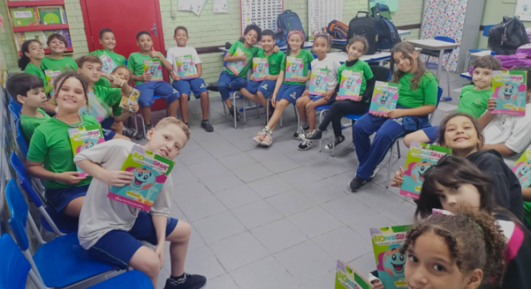 #paratodosverem: a foto mostra uma sala de aula com um grupo de crianças uniformizadas sentadas em círculo. Elas estão sorrindo e segurando o mesmo livro, intitulado "Biodiversidade", com capa colorida e ilustrada com um desenho do planeta terra. Ao fundo, há uma parede pintada de verde claro com cartazes educativos, além de mochilas sobre as mesas e uma estante com livros.