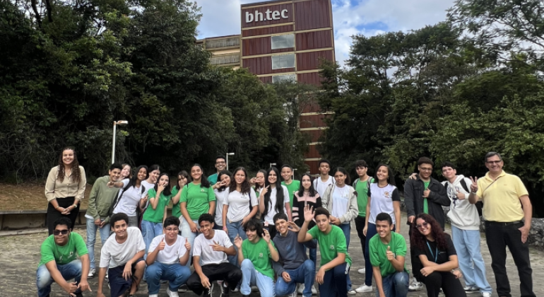 #paratodosverem: na imagem, grupo de estudantes e professores(as) posa para foto em área externa, com vegetação ao fundo e um prédio com a inscrição “bh.tec” ao centro da imagem. Os(As) estudantes demonstram animação, com gestos, acenos e sorrisos para a câmera. O céu está parcialmente nublado.