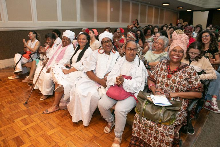 Feira e atividades culturais gratuitas marcam o encerramento do Julho das Pretas neste sábado (25) Auditório com dezenas de pessoas sentadas acompanhando atentamente uma atividade. Em primeiro plano, destacam-se duas mulheres negras: uma veste blusa vermelha de alças, turbante estampado em tons de vermelho, amarelo e preto, colares prateados e brincos, enquanto a outra usa vestido laranja, casaco claro, óculos de armação transparente e brincos de argola, apoiando o rosto na mão. Ao redor, há um público diverso, formado por mulheres e homens de diferentes idades e tons de pele.