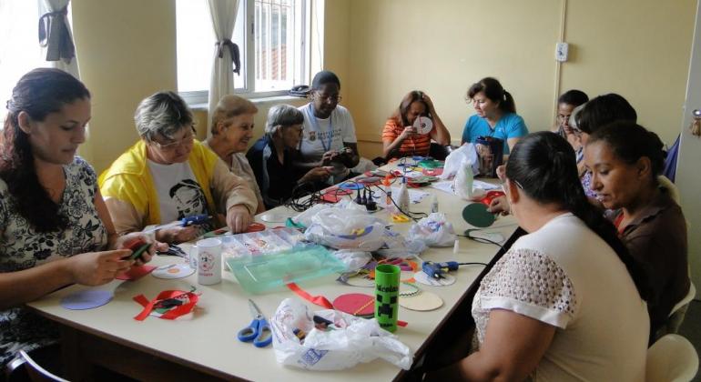 Dez mulheres, em torno de uma mesa, fazendo artesanato. 