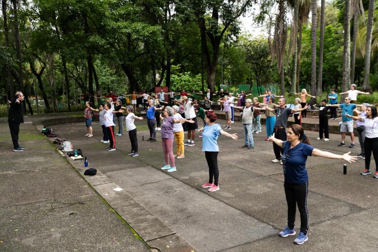A foto mostra um grupo de pessoas reunidas em um parque, fazendo aula de Lian Gong, que é uma prática corporal chinesa. 
