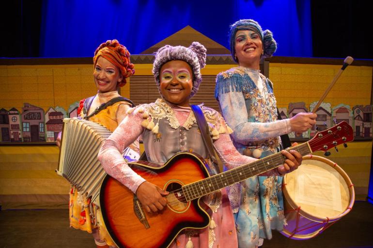 Teatro Marília recebe neste final de semana espetáculo que celebra a arte dos mamulengos Três atrizes do espetáculo "Mamulenga", do Grupo Girino, sorriem no palco segurando instrumentos musicais. A da esquerda toca acordeão, a do centro toca violão e a da direita carrega uma zabumba. Elas usam figurinos coloridos, maquiagem artística e cabelos feitos de lã estilizados.