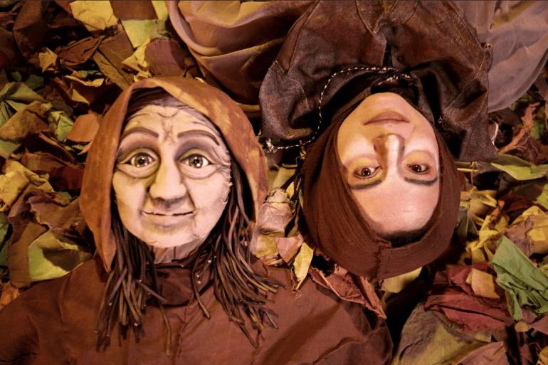 Montagem resgata saberes populares em apresentação no Teatro Marília Visão superior de duas figuras deitadas lado a lado sobre folhas coloridas: de um lado uma marionete de idosa com capuz marrom e olhos arregalados; do outro, uma mulher, com o rosto maquiado e cabeça coberta por um capuz marrom, olhando para cima.