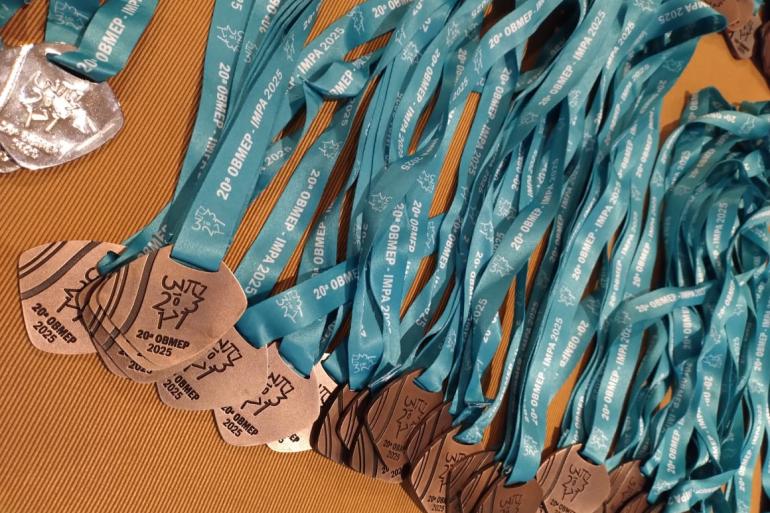 Alunos da Rede Municipal participam de cerimônia de entrega de medalhas da Olimpíada Brasileira de Matemática Medalhas da Olimpíada Brasileira de Matemática, com cordão azul claro, dispostas em uma mesa