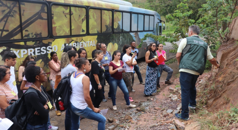 Treze pessoas, em frente a ao expresso ambiental, escutando instrutor da Secretaria Municipal de Meio Ambiente, em frente à mata.