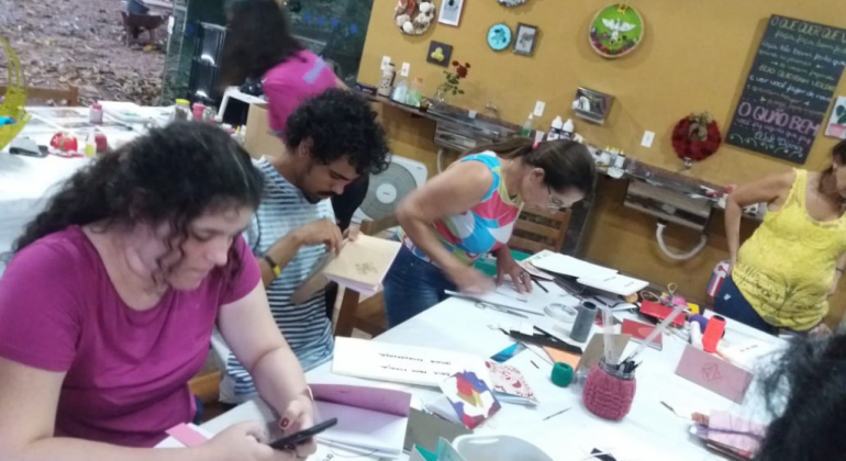 Prefeitura oferece, no mês de novembro, oficinas gratuitas de bordado mexicano e encadernação com costura artesanal na Pampulha. Foto: Divulgação PBH Quatro cidadãos ao redor de uma mesa com papéis fazendo oficina de encadernação com material reciclável.
