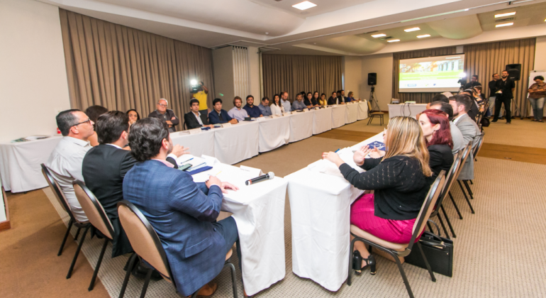 A escolha da capital mineira para construir as propostas foi feita em assembleia realizada em novembro em Recife (PE). Foto: Divulgação PBH Mesa em formato de "u" com trinta pessoas com telão ao fundo em uma sala.