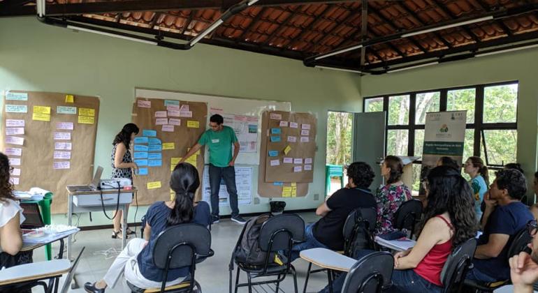 Homem, de pé, dá aula para mais de oito pessoas, sentadas, em sala de aula. 