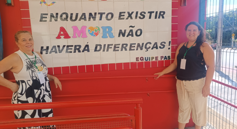 #paratodosverem: fotografia de duas mulheres sorrindo ao lado de um mural em parede vermelha. A frase no mural diz: "AUTISMO — Enquanto existir AMOR, não haverá diferenças!" As palavras "AUTISMO” e "AMOR" estão escritas com letras de cores variadas, e a letra "O" é representada por um coração com estampa de peças de quebra-cabeça, símbolo do autismo. O restante da frase está em preto.