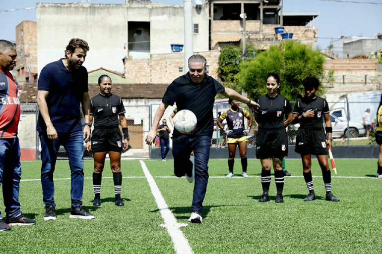 Homem branco,, prefeito Álvaro Damião, trajando calça jeans escura e camisa preta chuta uma bola de futebol branca em um campo de futebol. Ao lado dele um homem branco e três árbitras de futebol observam 