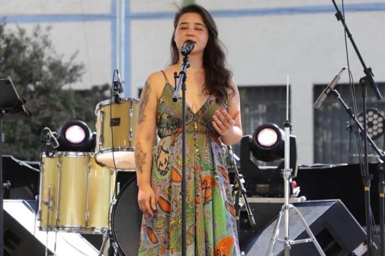 Cantora Natália Coutinho se apresenta em um palco ao ar livre, com os olhos fechados e expressão concentrada, enquanto canta ao microfone. Ao fundo, aparecem instrumentos musicais, equipamentos de som e refletores