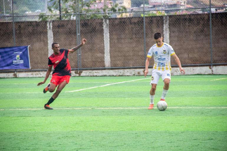 Copa Centenário chega à 25ª rodada com 14 jogos dos módulos A, B e C neste domingo (20) Dois jogadores disputam a bola em uma partida de futebol society, realizada em campo de grama sintética. Um atleta veste uniforme vermelho e preto, enquanto o outro usa uniforme branco e amarelo.