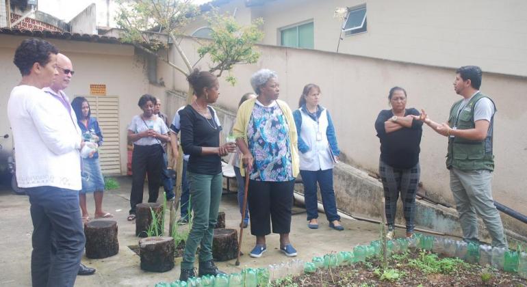 Hortas comunitárias nas unidades básicas de saúde de BH funcionam como integração comunitária e ação terapêutica. Foto: Mara Damasceno/PBH Nove pessoas próximas à horta comunitária do centro de saúde, durante o dia.