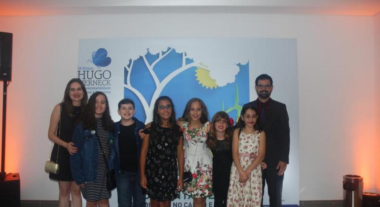 Foto de seis crianças e dois professores em frente a um banner do prêmio Hugo Werneck
