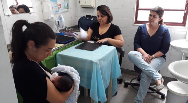 Imagem traz mulher amamentando bebê, de frente para outras duas mulheres