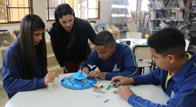 Três alunos de escola municipal reúnem objetos em cima da mesa, acompanhados por mulher.