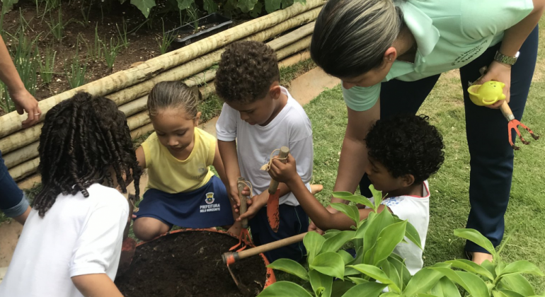 Quatro crianças plantam em recipiente redondo, acompanhadas por adulto, em local aberto, durante o dia. 