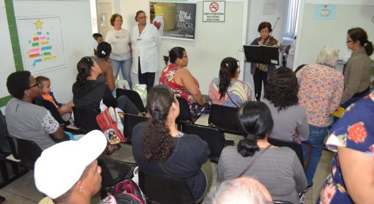 Foto: Camila Rodrigues Mais de dez pssoas assistem a mulher tocando violino em sala de centro de saúde.