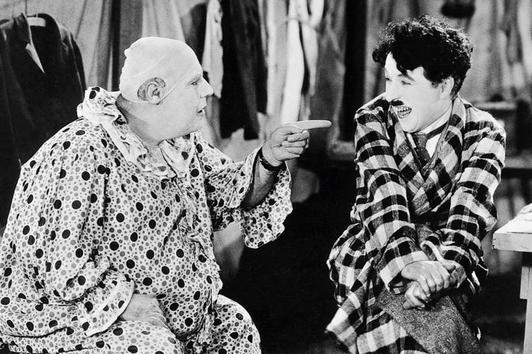 Cena de filme em preto e branco com Charlie Chaplin. A esquerda um personagem sentado com roupas de palhaço, sem peruca e apontando o dedo para a direita em direção ao personagem de Chaplin, que veste uma roupa xadrez e está sentado com as pernas cruzadas. 