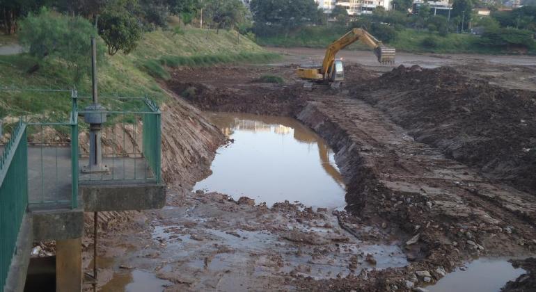 Ação é uma etapa importante para prevenção de enchentes do Córrego Leitão e contribui para diminuição de problemas na av. Prudente de Morais. Foto: Antonio Rodrigues/PBH Escavadeira retira terra da Barragem Santa Lúcia. Ao fundo, árvore e prédios, durante o dia.