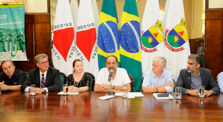 Prefeito e secretariado assinam convênio com representantes da Caixa Econômica Federal, todos sentados na mesa do Salão Nobre da PBH. 