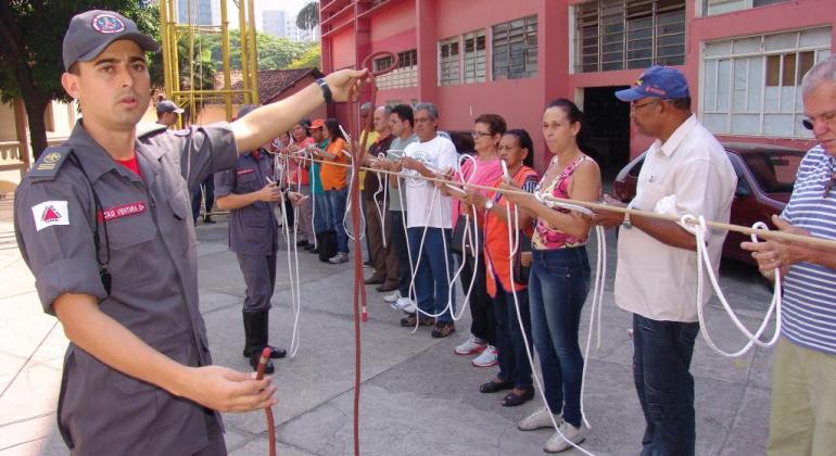 Bombeiro orienta mais de dez voluntários, de pé, a dar nós. 