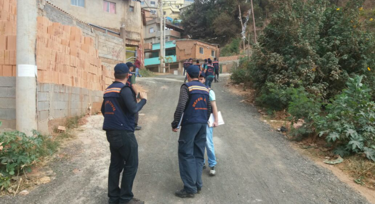 Dois técnicos da Defesa Civil acompanham cidadão em vila, mais à frente, outros dois técnicos caminham. Foto ilustrativa.