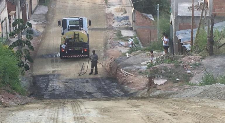 Rua de terra em início de pavimentação com caminhão ao fundo, durante o dia. 