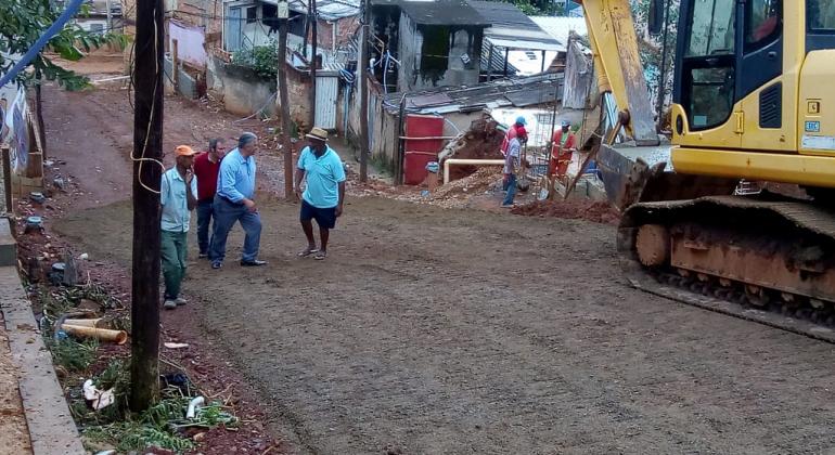 Rua General Osorio, com um declive, sendo pavimentada por máquina. Ao fundo, três pessoas contemplam a pavimentação. 