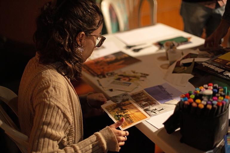 “Museus Centro” conecta patrimônio, criação e temas contemporâneos na programação de agosto Uma mulher jovem, de cabelos castanhos cacheados presos parcialmente para trás, está sentada à mesa participando de uma atividade de colagem. Ela usa óculos, um casaco de tricô claro e segura fotografias impressas enquanto observa atentamente os materiais à sua frente. Sobre a mesa há revistas, recortes, canetas coloridas, tesoura e outros materiais de papelaria organizados para a oficina. A iluminação é baixa, com luz quente direcionada para a mesa, criando um ambiente acolhedor.