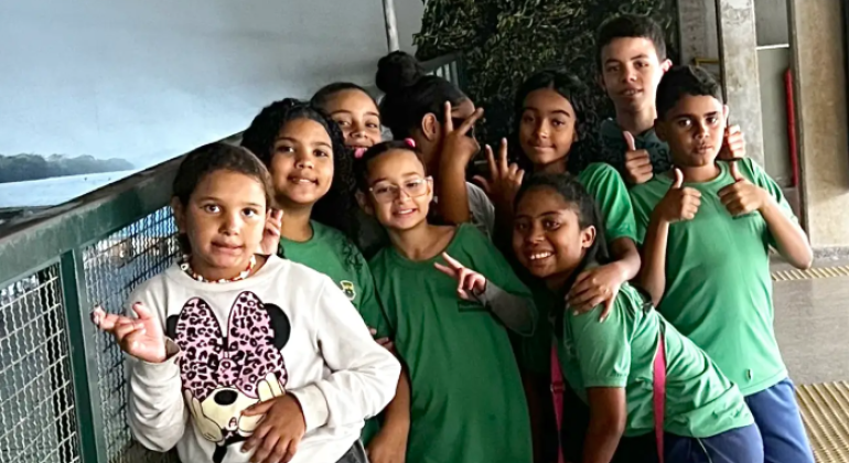 #paratodosverem: a foto mostra um grupo de estudantes em um ambiente interno. A maioria está vestida com o uniforme escolar, composto por camiseta verde e bermuda azul. À esquerda, uma aluna veste blusa branca com estampa da Minnie Mouse e calça preta. O local tem piso de granito e uma divisória metálica à esquerda. 