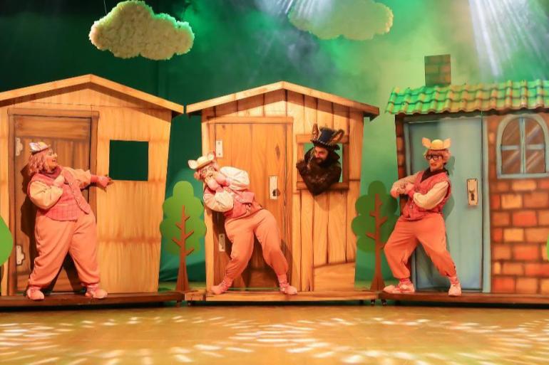 Teatro Francisco Nunes recebe nova montagem da renomada história “Os 3 Porquinhos” No palco, três atores vestidos de porquinhos com roupas rosa e chapéus estão posicionados em frente a três pequenas casas de cenografia — uma de palha, uma de madeira e uma de tijolos. O Lobo Mau, caracterizado de escuro com garras, espia por uma janela da casa do meio enquanto os porquinhos fazem frases expressivas de surpresa e alerta.