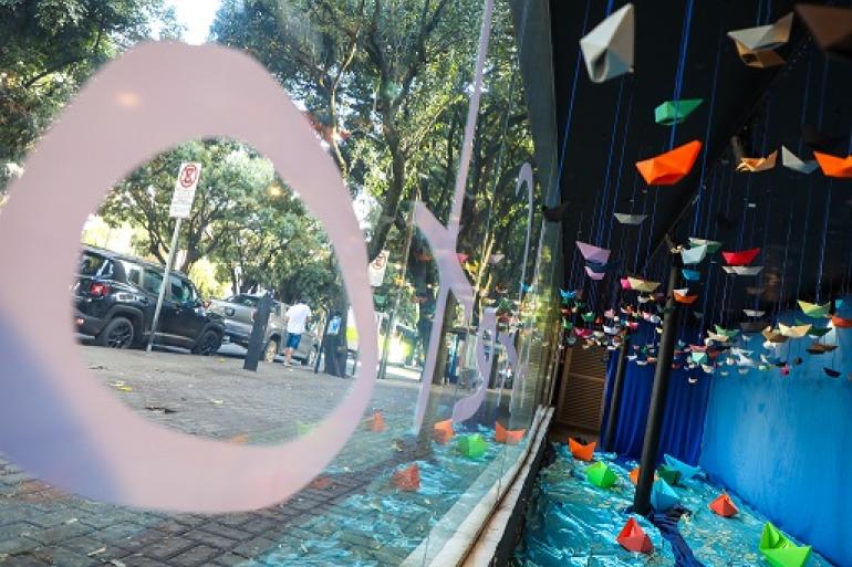 Vitrine do Teatro Marília recebe a exposição “O Trajeto do Afeto”, de Paloma Parentoni Vitrine com barquinhos de papel pendurados de várias cores