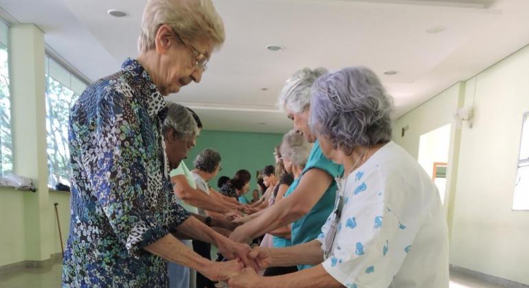 Grupo de Dança Sênior da Prefeitura de Belo Horizonte promove bem-estar e melhora autoestima da pessoa idosa. Foto: Andréa Moreira/PBH Duas idosas dançam de mãos dadas, olhando uma para a outra, no fundo, mais de oito pares de idosos repetem o gesto.