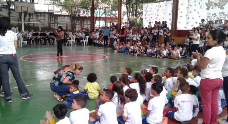 Mais de duzentos alunos em ginásio da Escola Municipal Anne Frank. Ao centro, mulher fala a todos. 