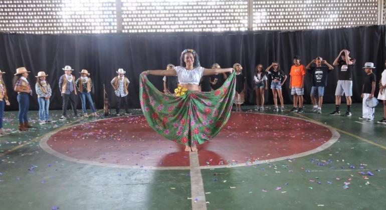 Estudante com saia florida e outros estudantes ao fundo faz apresentação de dança. 