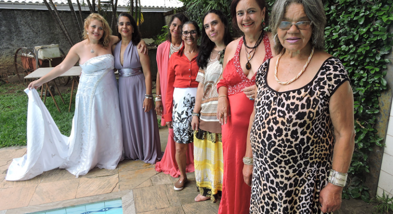 Mulheres posam para foto usando vestidos longos, ao lado de uma piscina