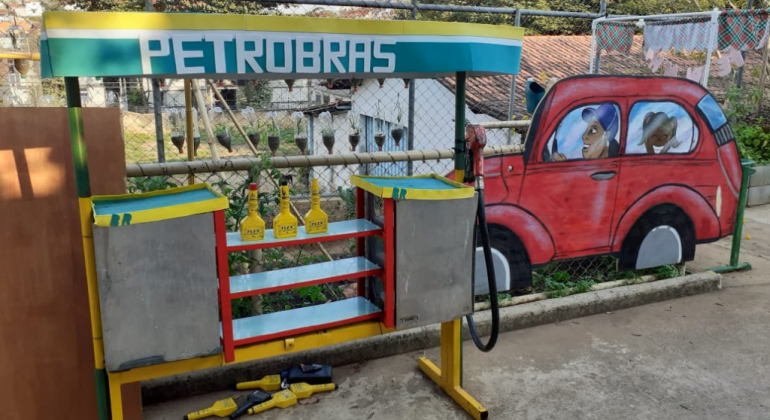 Maquete colorida de posto de gasolina, com desenho de carro ao lado, durante o dia.
