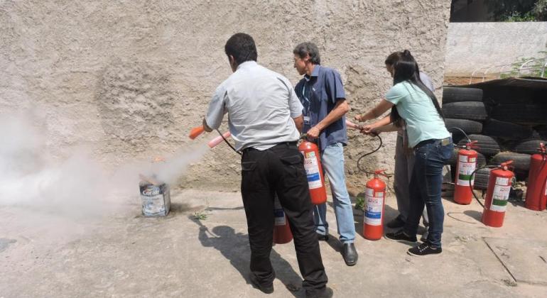 Três pessoas aprendem a manusear extintores de incêndio, apagando uma lata de tinta com fogo dentro, em pátio. 