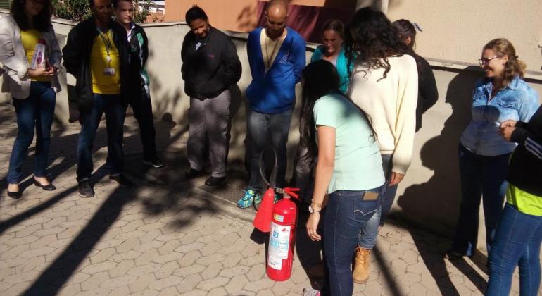 Sete pessoas observam uma mulher manejar o extintor de incêndio durante o dia. 