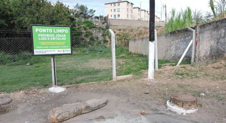 Calçada e local gramado com grade, limpos, com uma placa com os dizeres: "Ponto limpo de entulho", durante o dia. 