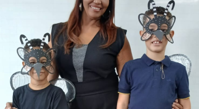 #paratodosverem: a foto mostra uma professora entre dois estudantes. As crianças usam máscaras e asas representando o mosquito da dengue. Todos(as) estão sorrindo. O fundo é um quadro branco e parte de uma parede de azulejos brancos. 