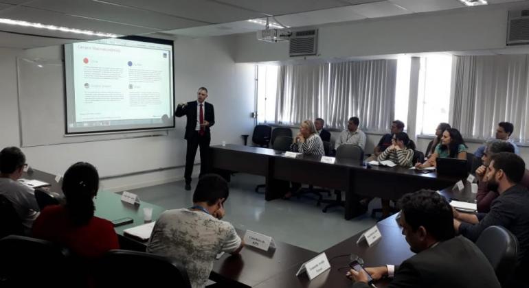 Homem de terno palestra em frente à mesa em forma de "u" com cerca de 13 pessoas. Ao fundo, painel com projeção de imagens.