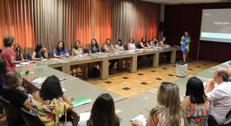 Dezessete pessoas reunidas em mesa em formato de "u", acompanhando palestra de mulher, ao lado de telão, em sala da PBH. 