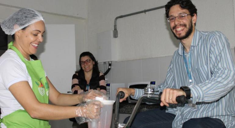 Homem faz teste ergométrico enquanto mulher prepara suco no liquidificador, em ambiente fechado. 
